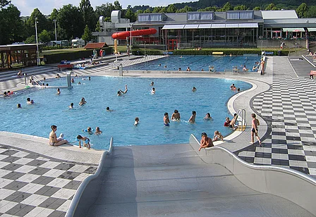 Familienfreundliches Schwimmbad mit mehreren Pools, Spielmöglichkeiten und Sonnenbadeträumen. Viele Badegäste genießen die Sonne und das Wasser.