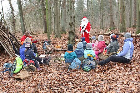 Gruppe von Kindern sitzen im Wald. Eine Frau spielt Gitarre und der Nikolaus steht vor den Kindern.
