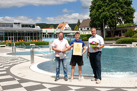 Noah Böhme hat seine Meisterprüfung bestanden. Geschäftsführer Bernd Cranen und Teamleiter Bäder Bernd Kunert gratulieren Herrn Böhme vor dem Nichtschwimmerbecken im Einbecker Freibad und überreichen einen Blumenstrauß und ein Präsent. Herr Böhme hält seine Meisterukunde vor sich.